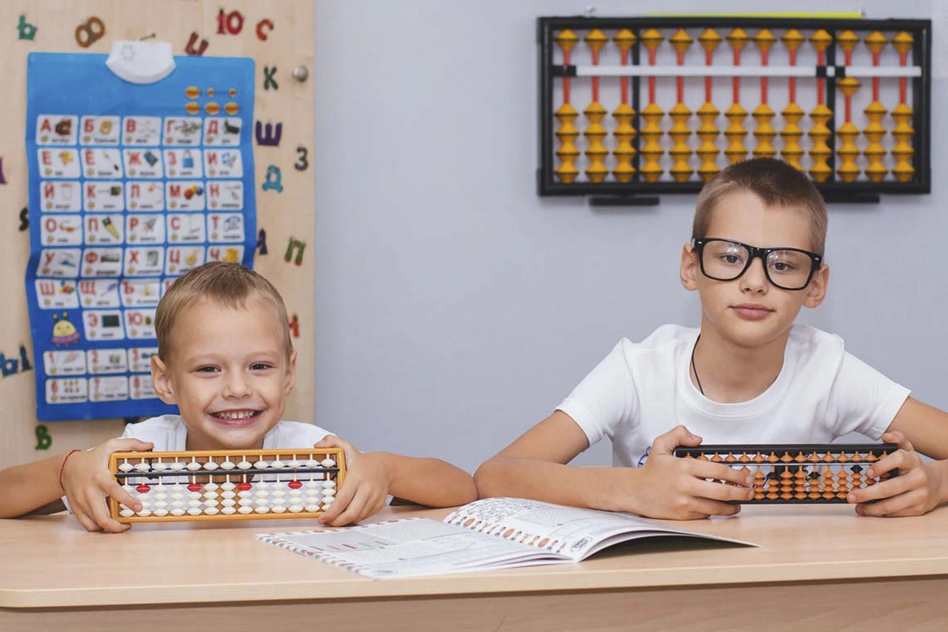 Dwóch chłopców w szkolnej ławce trzymających liczydła. Chłopiec po prawej stronie nosi czarne, prostokątne okulary korekcyjne.